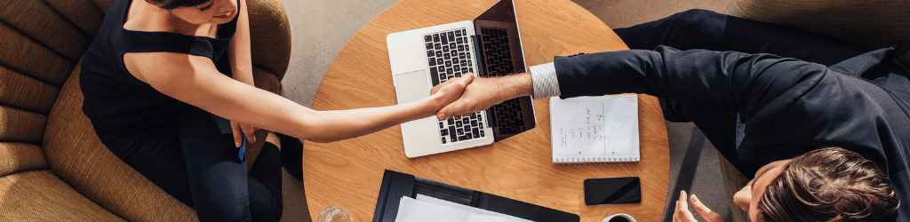 Two people shaking hands over a table with a laptop, tablet, notebook, and cellphone.