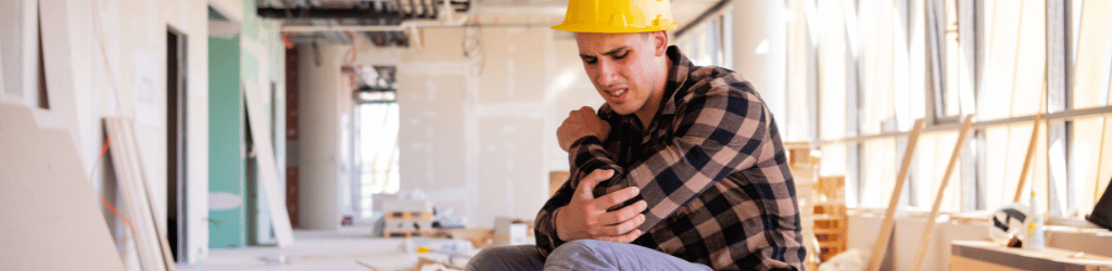 Man wearing a flannel, and yellow hard hat, clutching elbow in pain.