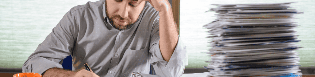 Man paying bills in home office with towering stack of bills on desk.